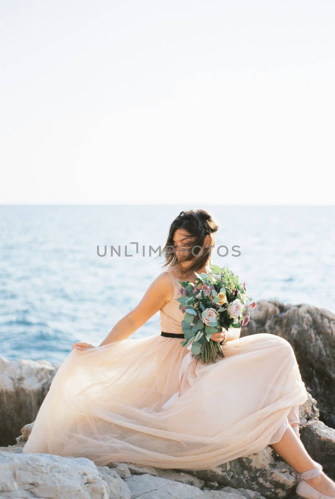 Bride with hair fluttering in the wind adjusts her dress sitting on the rocks by the sea. High quality photo