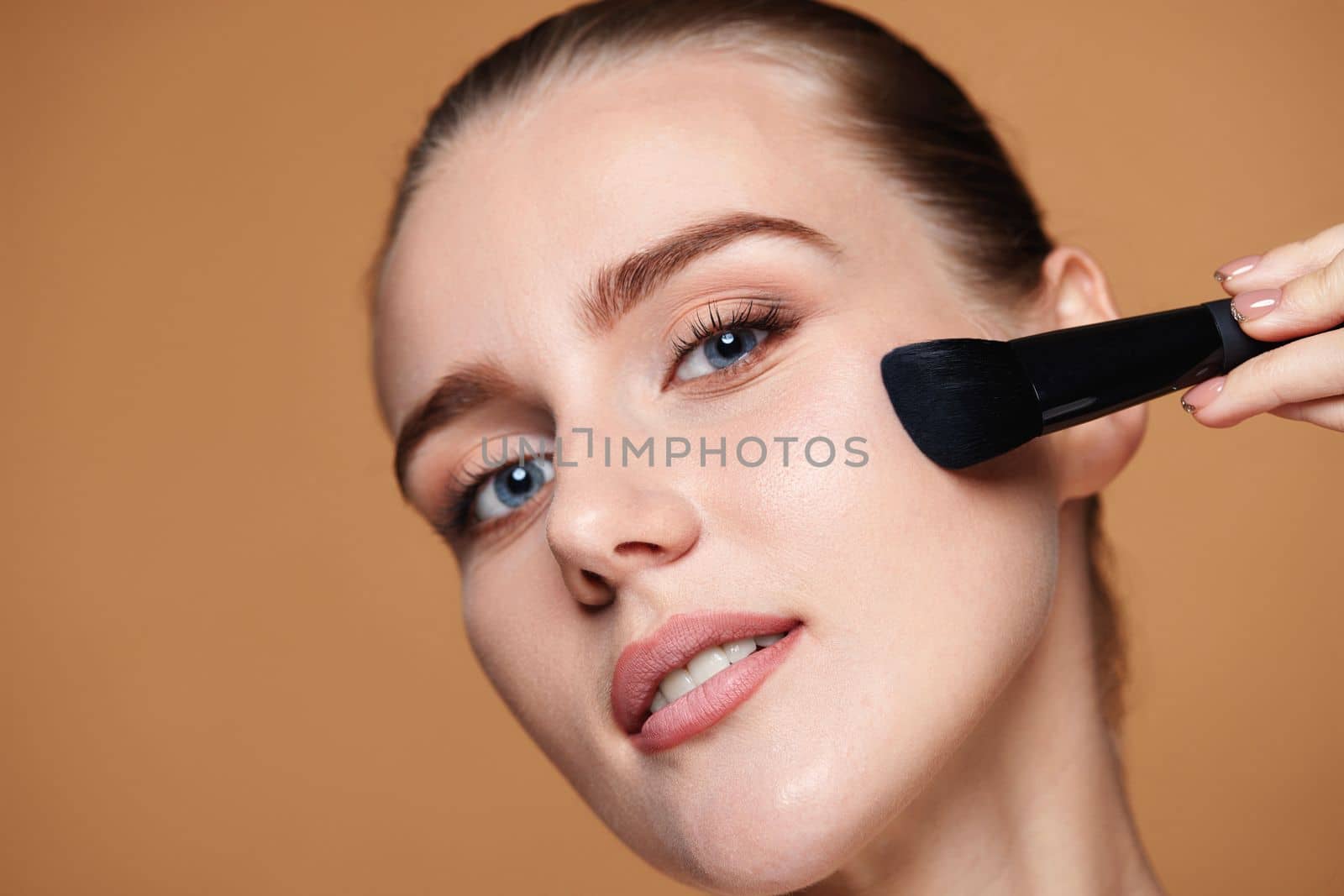 young woman applying blush on cheekbone with make-up brush on beige background. contouring