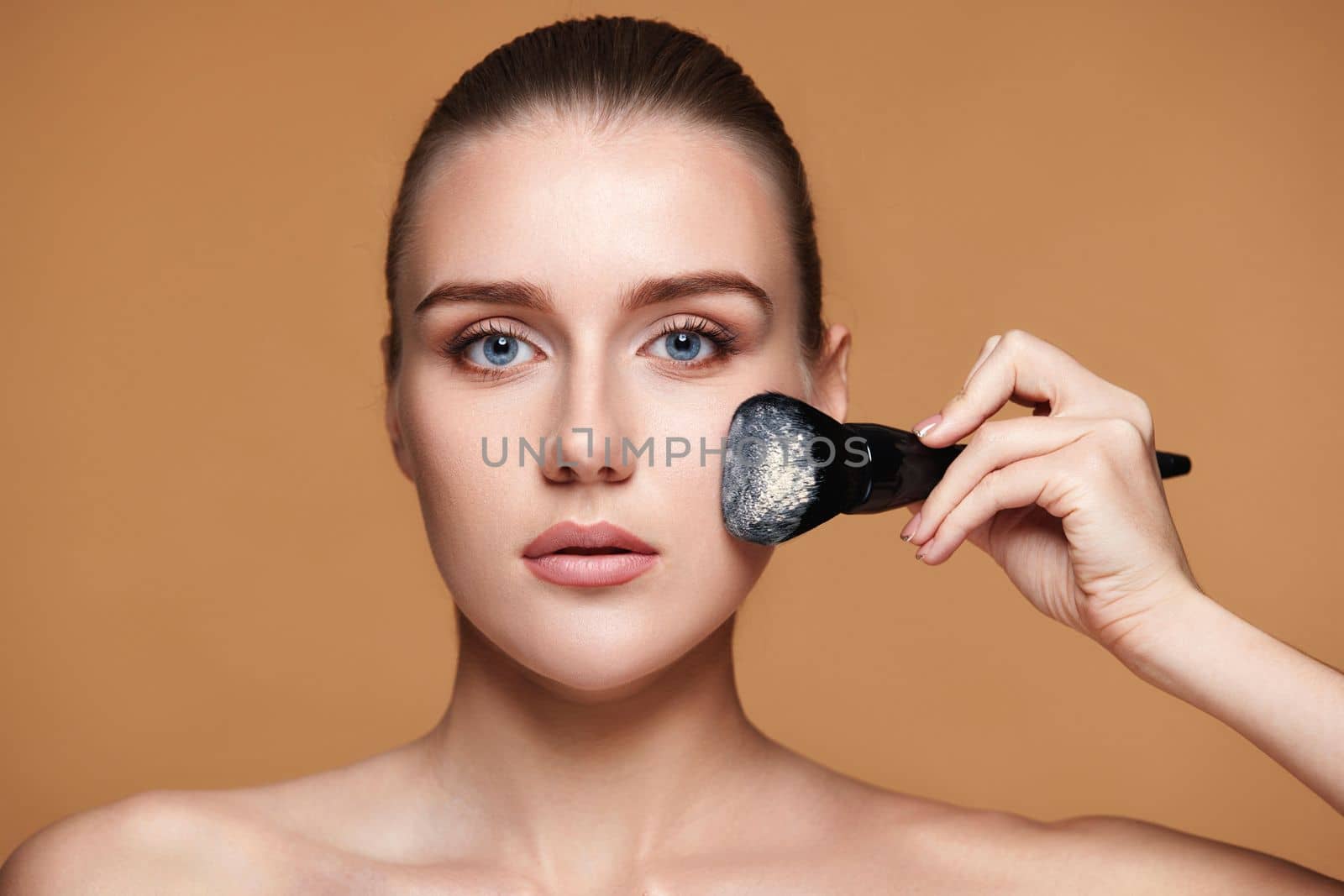 pretty young woman applying powder with professional make-up brush on beige background