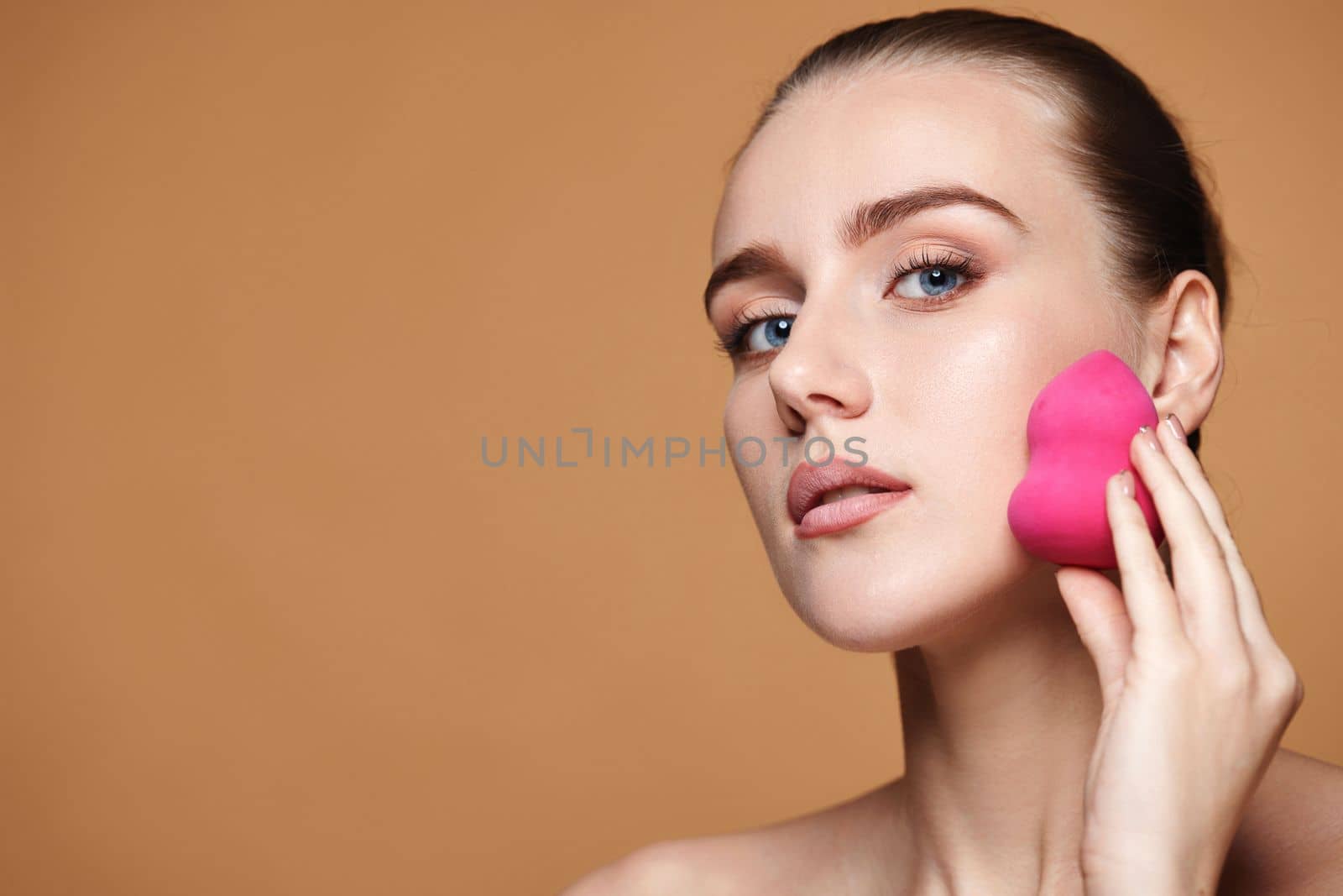 attractive young woman using pink cosmetic sponges on beige background. copy space