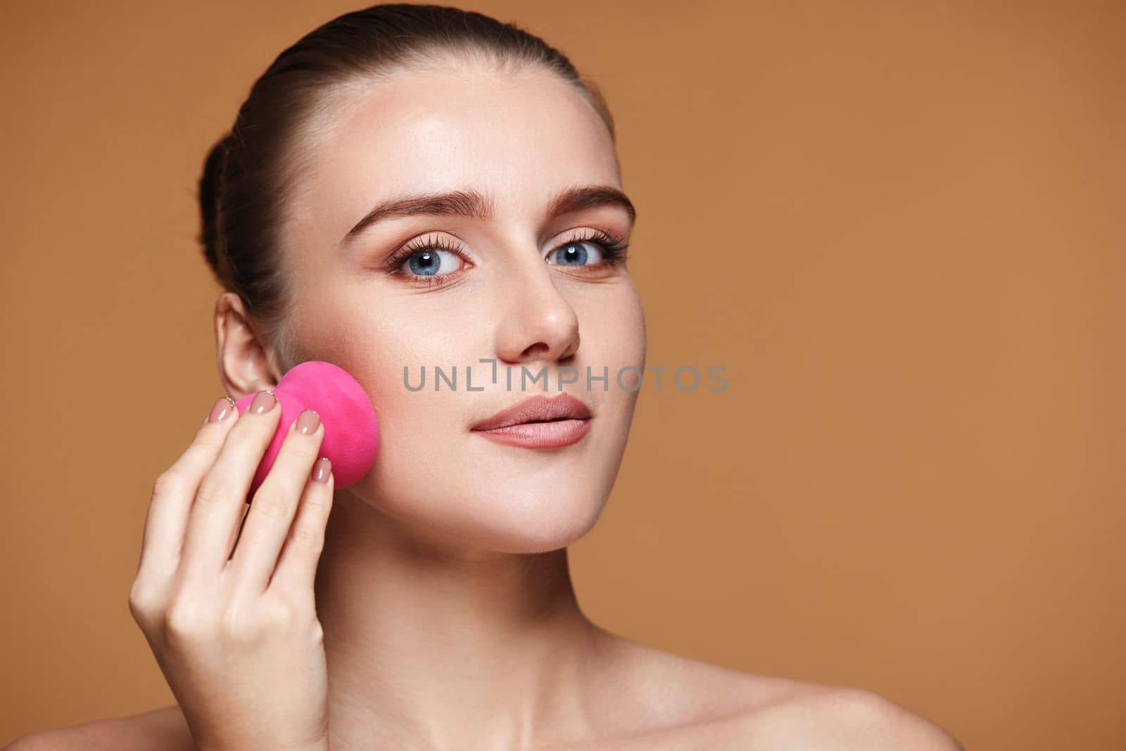 smiling woman applying foundation on skin with beauty blender on beige background