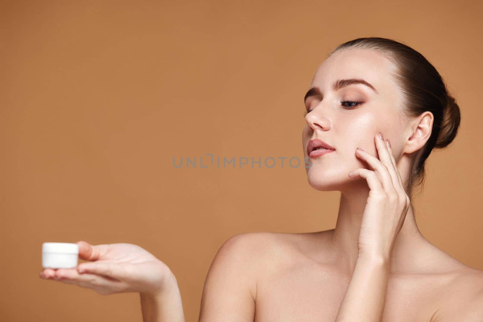 charming woman applying skin care cream on her face and holding jar of cosmetic product on beige background. copy space