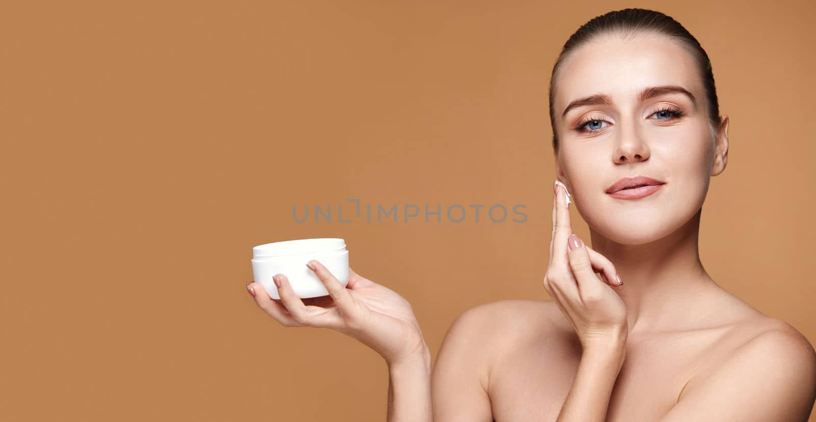 beautiful woman applying skin care cream on her face and holding jar of cosmetic product on beige background. copy space