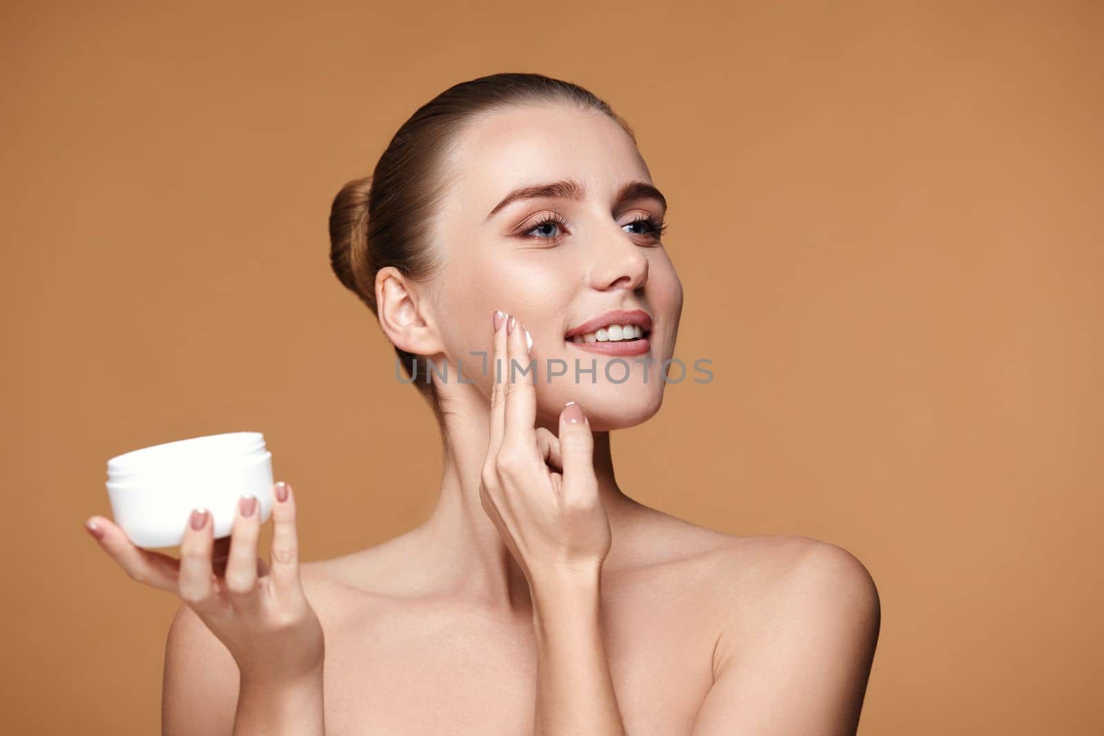 beautiful smiling young woman applying skin care cream on her face on beige background