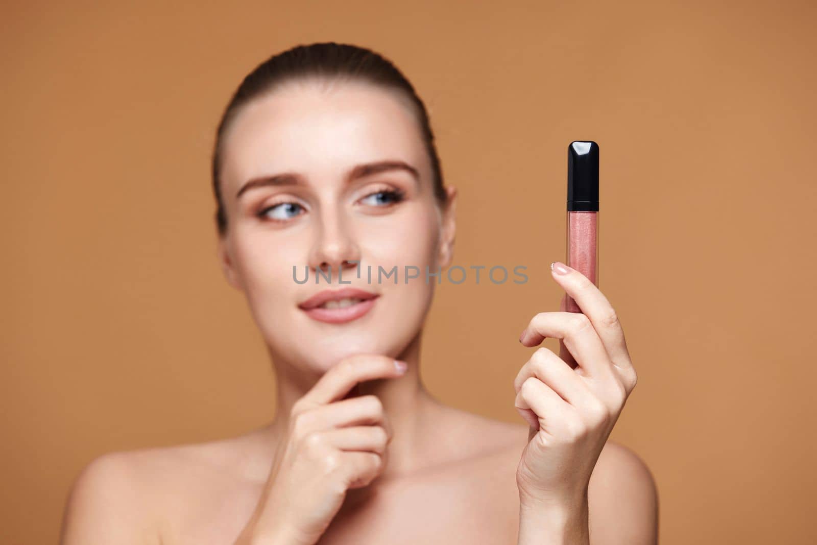 thinking smiling woman look away at nude lip gloss isolated on beige pastel background. focus on lip gloss