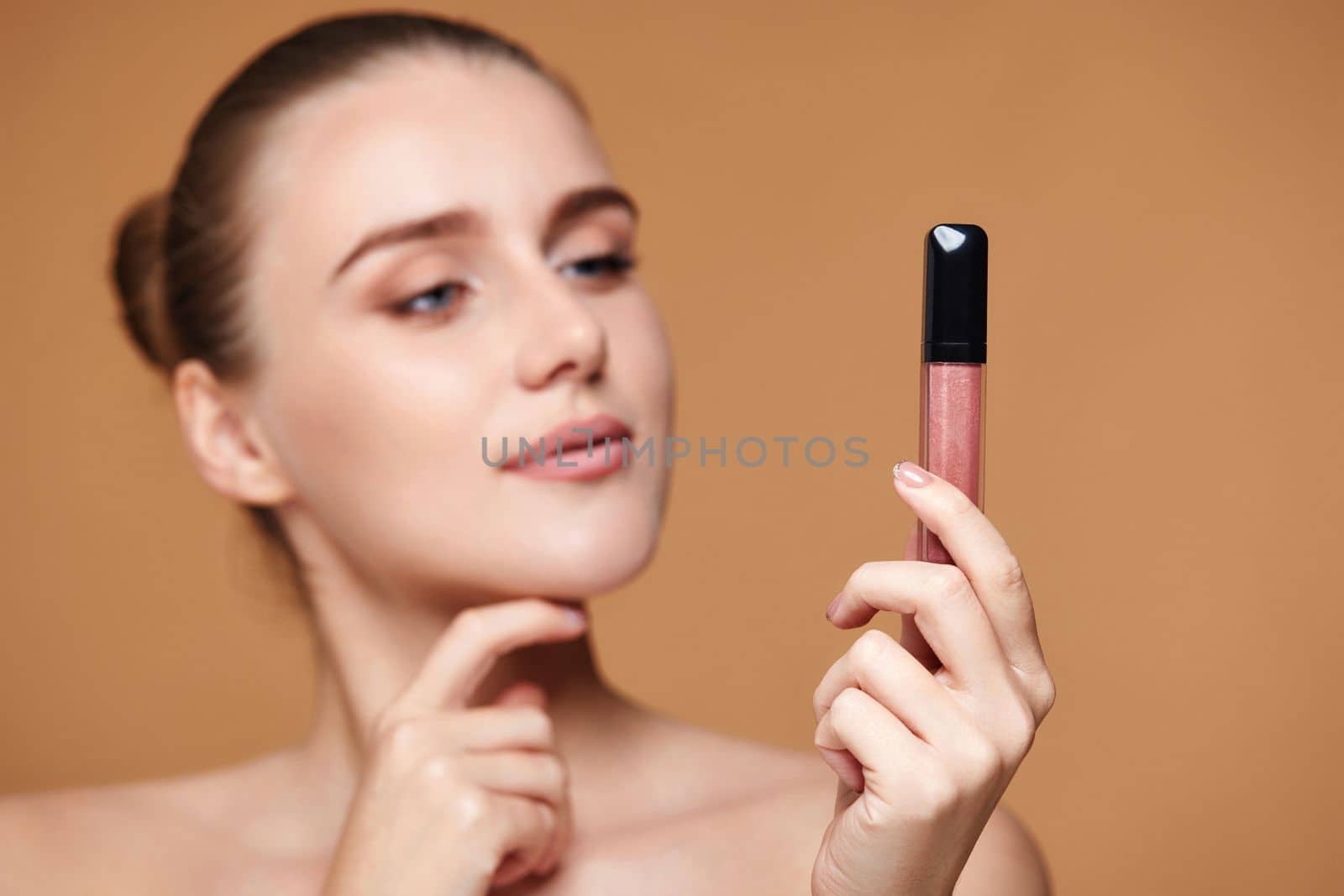 beautiful thinking woman look away at nude lip gloss isolated on beige pastel background. focus on lip gloss