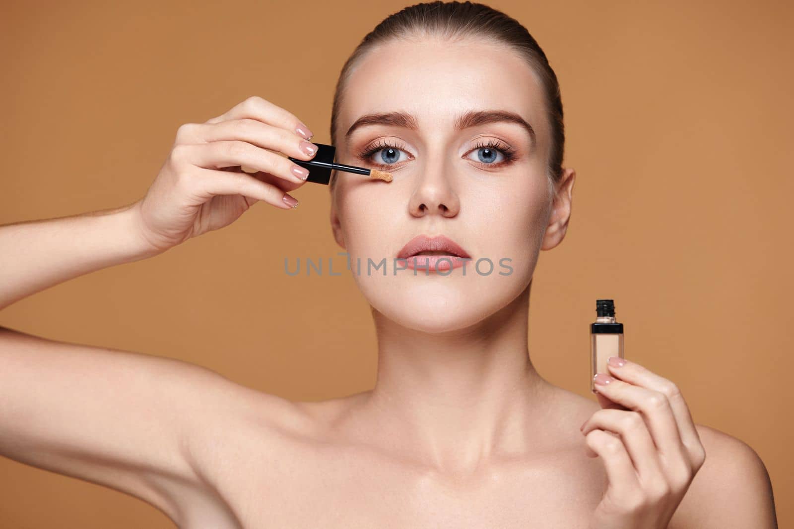 portrait of beautiful woman applying concealer under her eyes on beige background