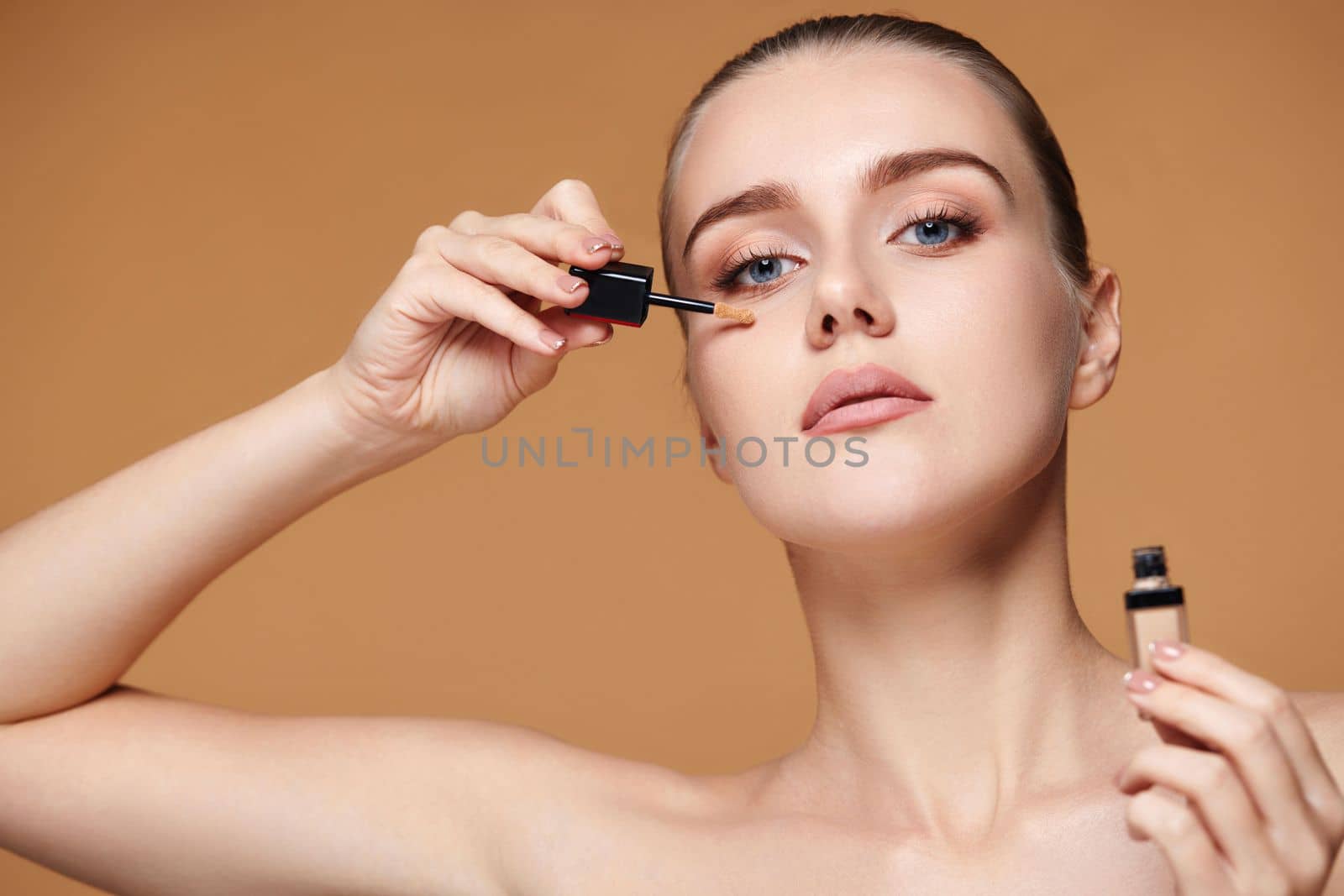 portrait of beautiful woman applying concealer under her eyes on beige background