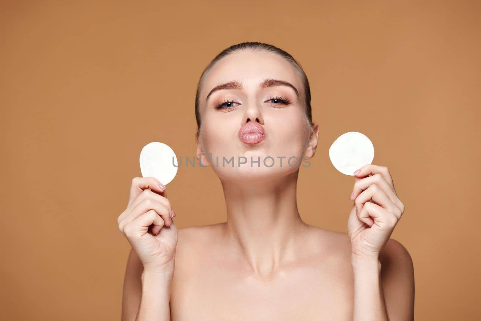 astonished beautiful woman holding two white cotton pad with micellar water on beige background
