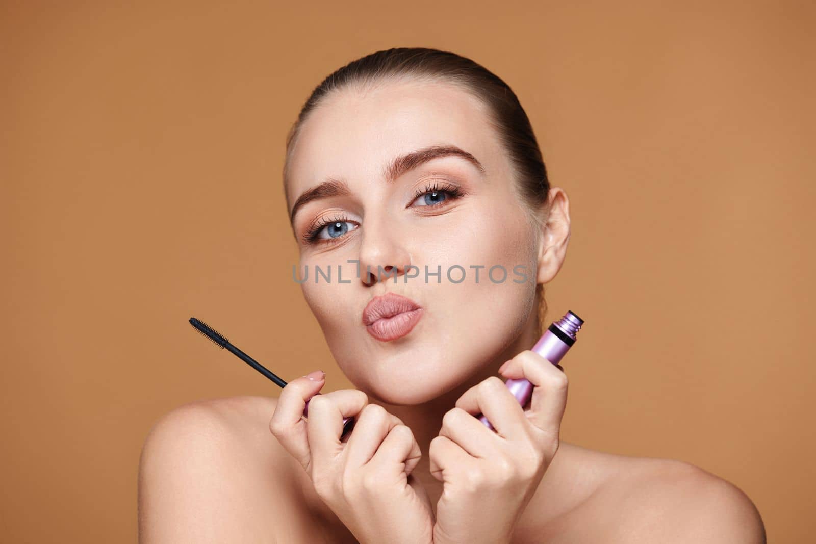 attractive young woman holding mascara and sending air kiss on beige background