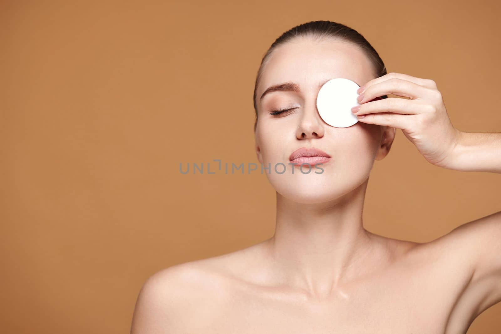 beautiful woman with naked shoulders close one eye with white cotton pad on beige background