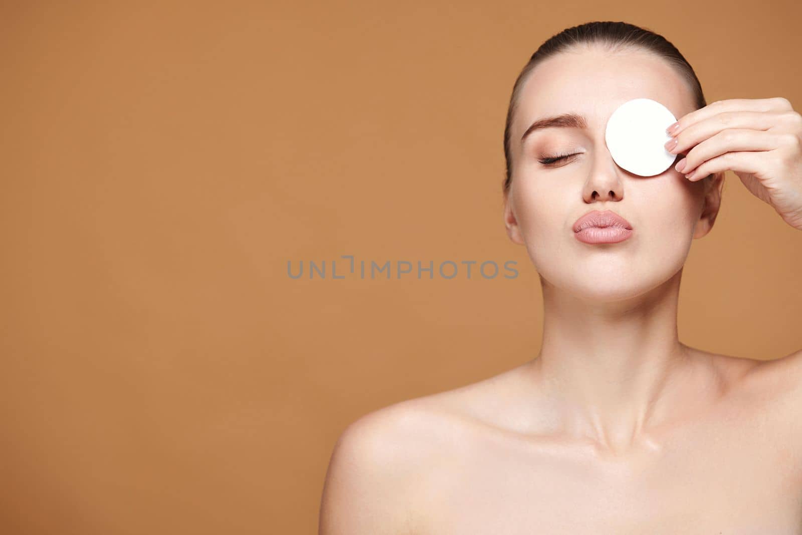 beautiful woman with naked shoulders close one eye with white cotton pad on beige background