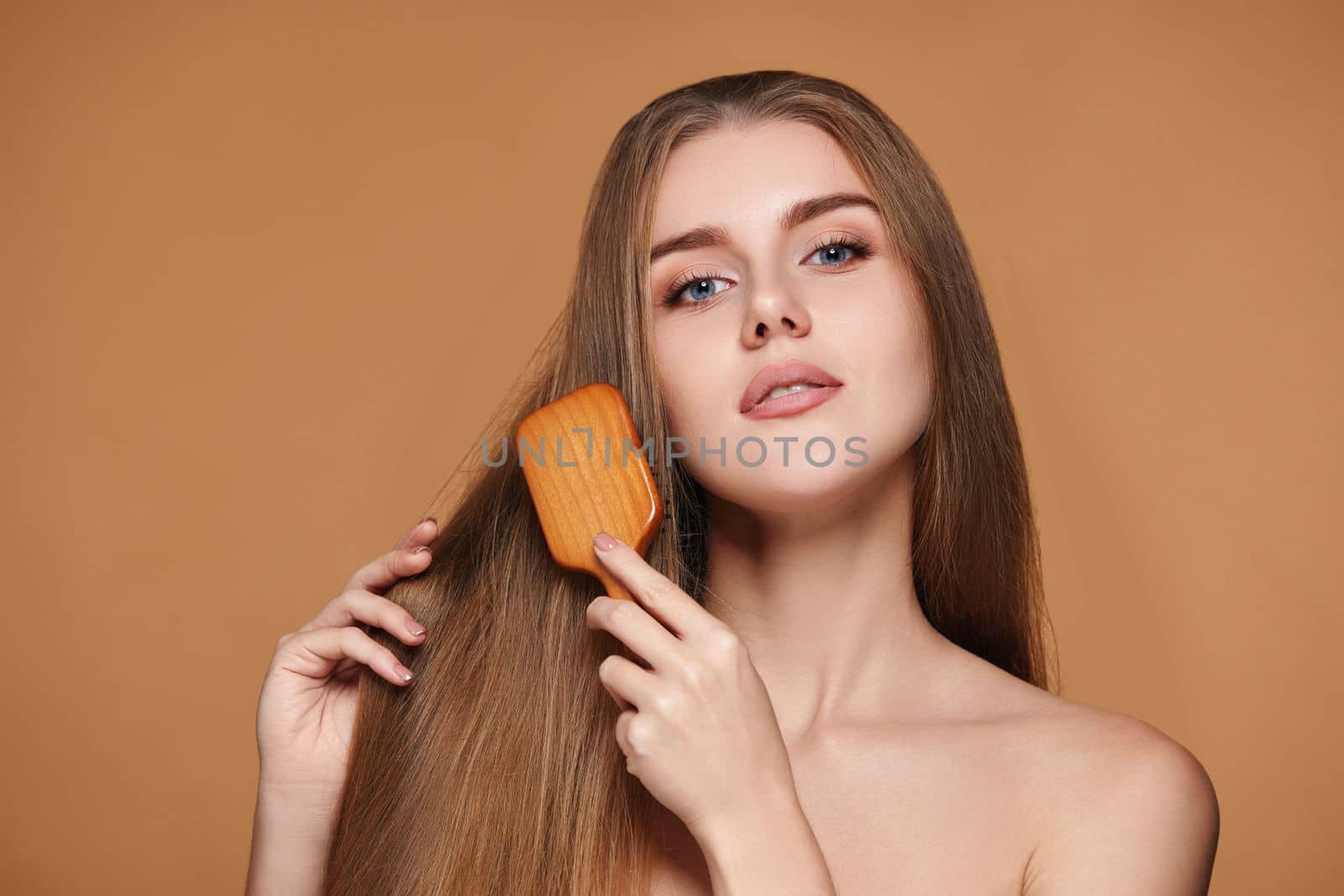 Beautiful woman looks at the camera and combs her long natural hair with wooden brush on beige background