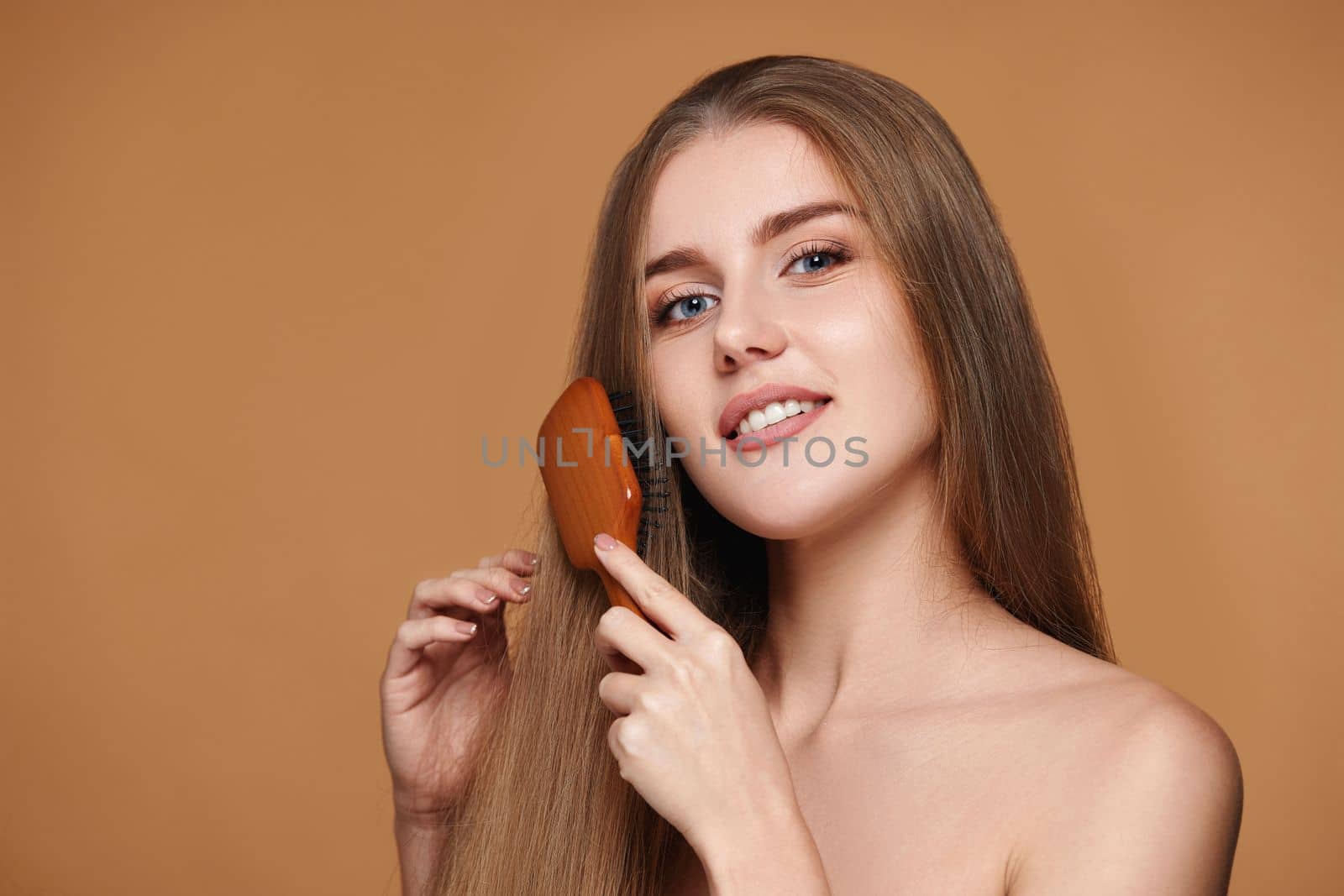 Beautiful smiling brunette woman looks at the camera and combs her long natural hair with wooden brush isolated on beige background
