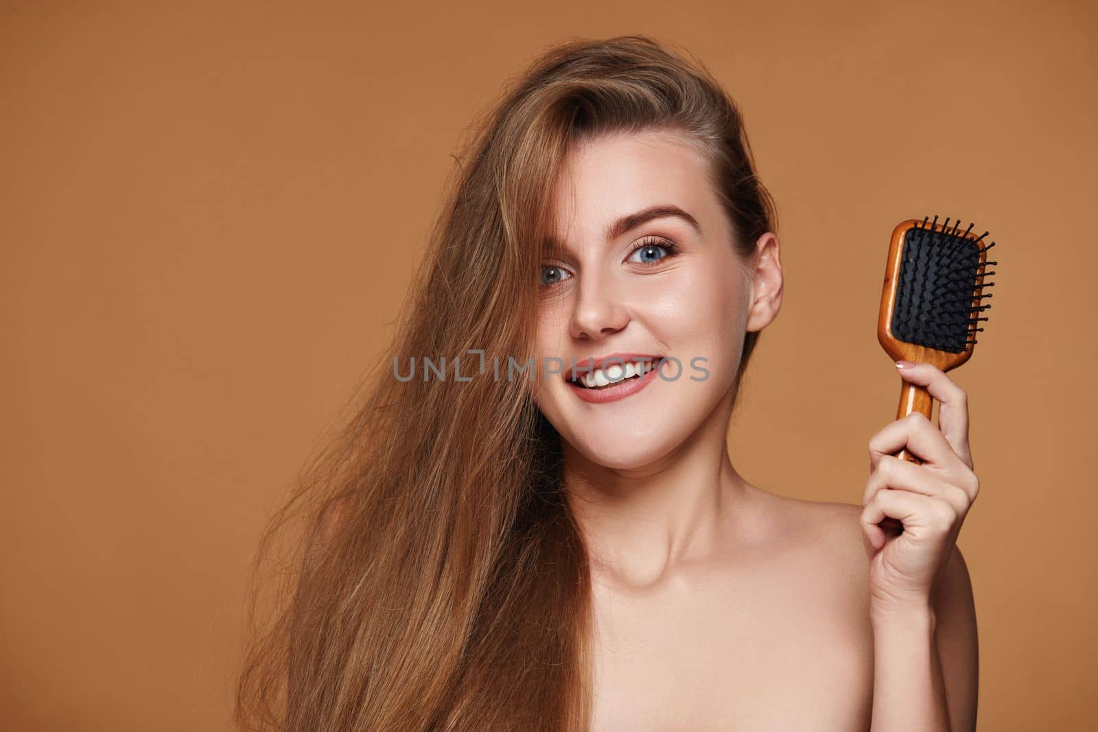 Beautiful hapy brunette woman showing wooden brush for healthy long hair on beige background