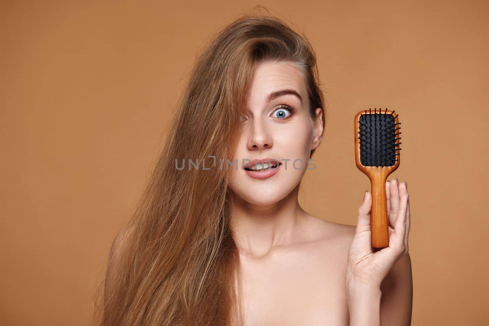 surprised beautiful brunette woman showing wooden brush for healthy long hair on beige background