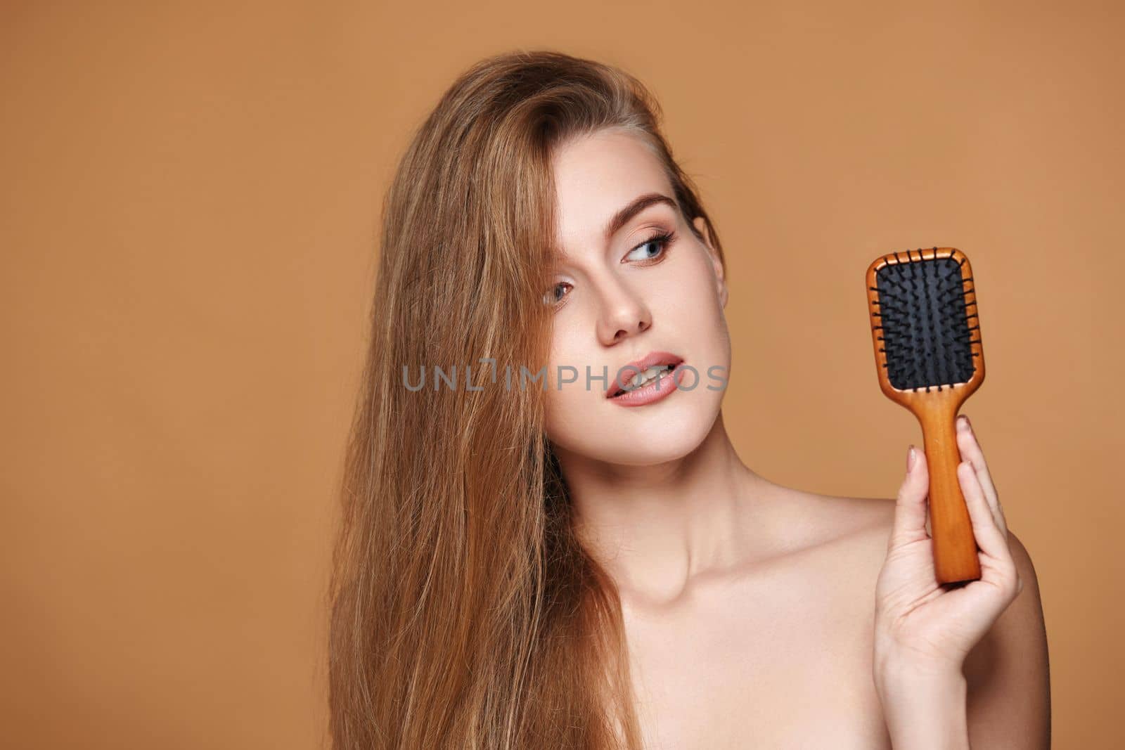 Beautiful brunette woman showing wooden brush and recommend haircare product for healthy long hair on beige background