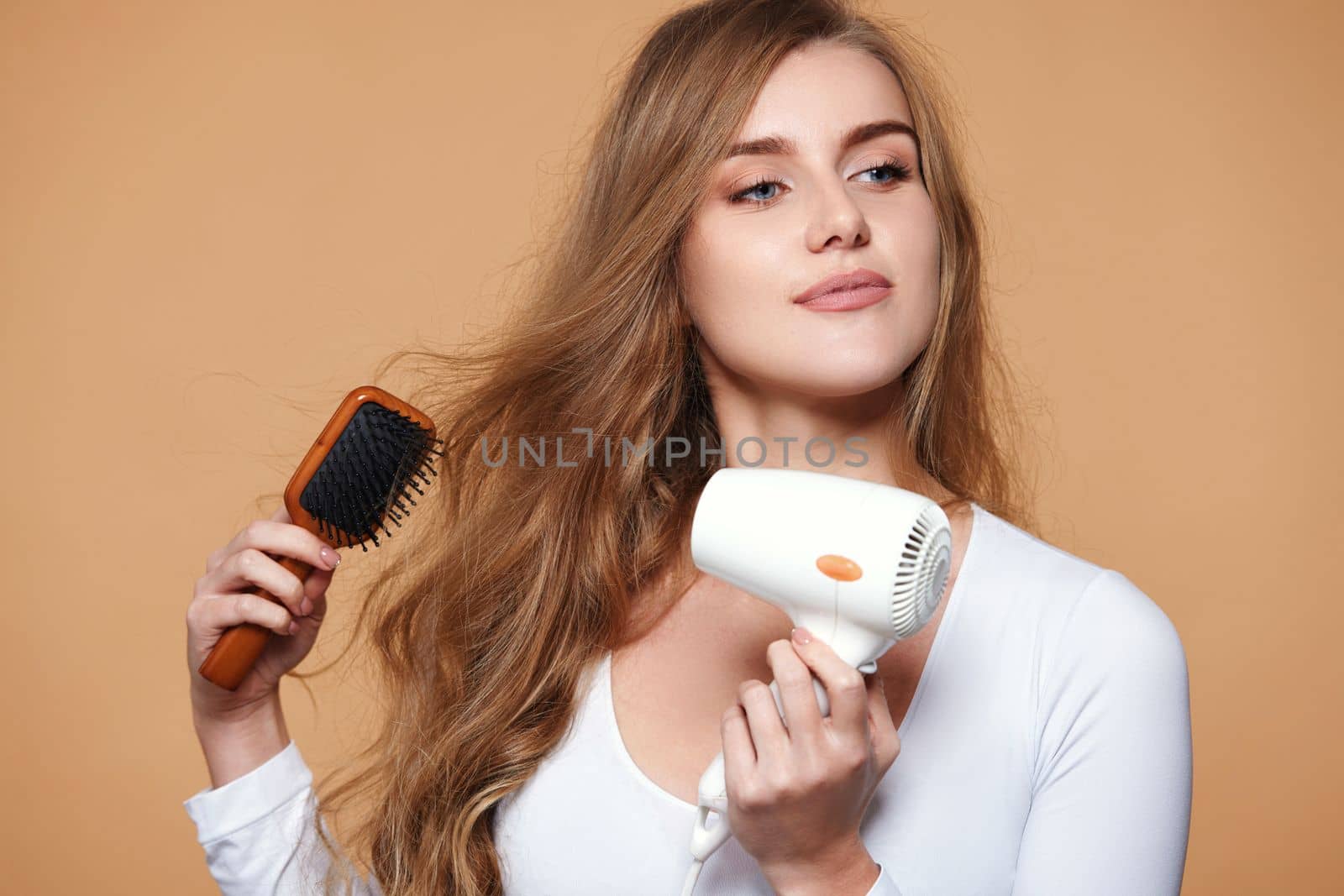 beautiful young happy woman drying strong healthy curly hair with dryer on beige background
