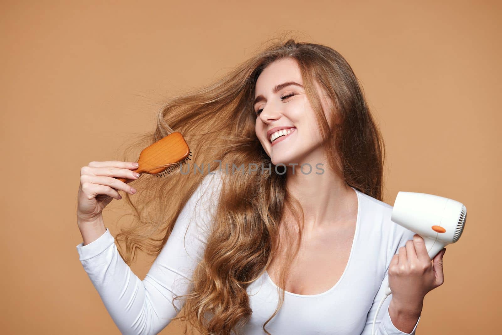 Portrait of attractive cheerful lady drying strong healthy curly hair using white hair dryer on beige background