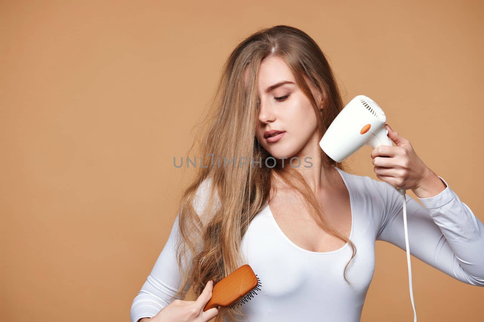 lovely woman with strong healthy curly hair using white hair dryer on beige background