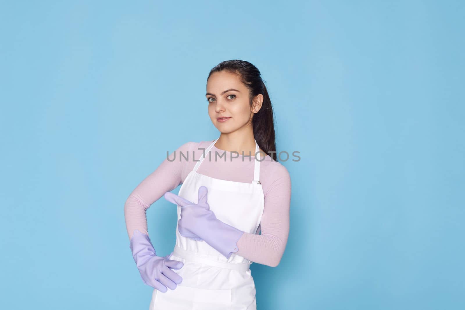 beautiful caucasian woman in violet rubber gloves pointing fingers aside at copy space on blue background. cleaning