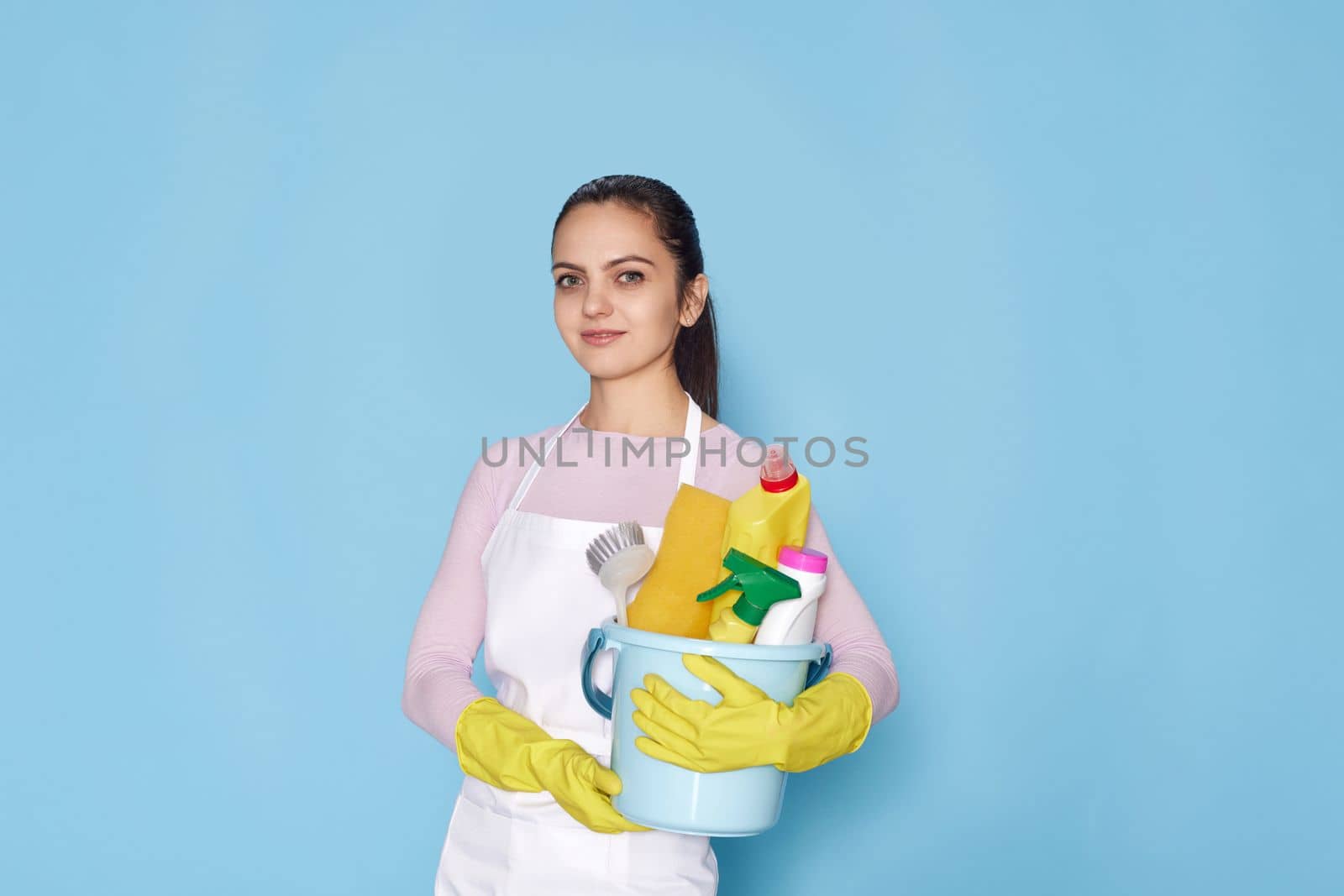 smiling caucasian woman in gloves holding bucket with cleaning supplies isolated on blue background.