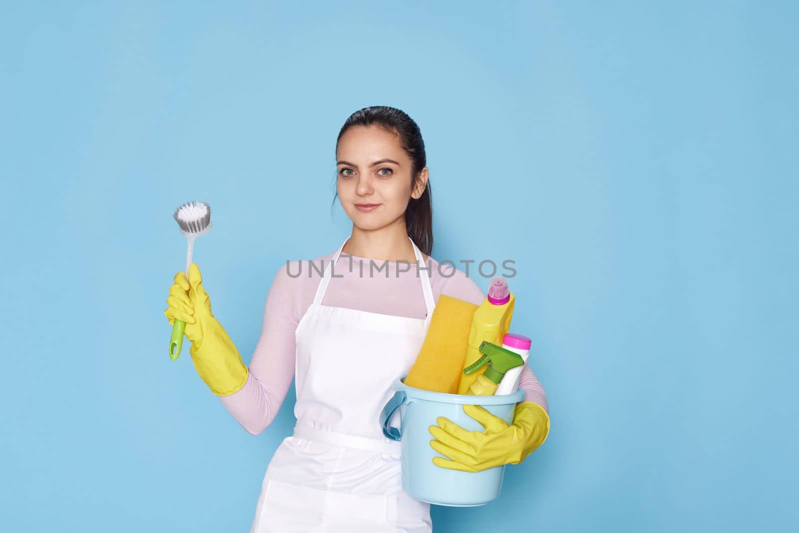 pretty woman in rubber gloves and cleaner apron holding bucket of detergents and toilet brush on blue background.