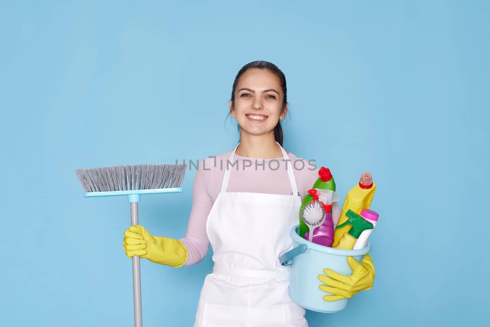 happy woman in yellow rubber gloves and cleaner apron holding bucket of detergents and broom on blue background.