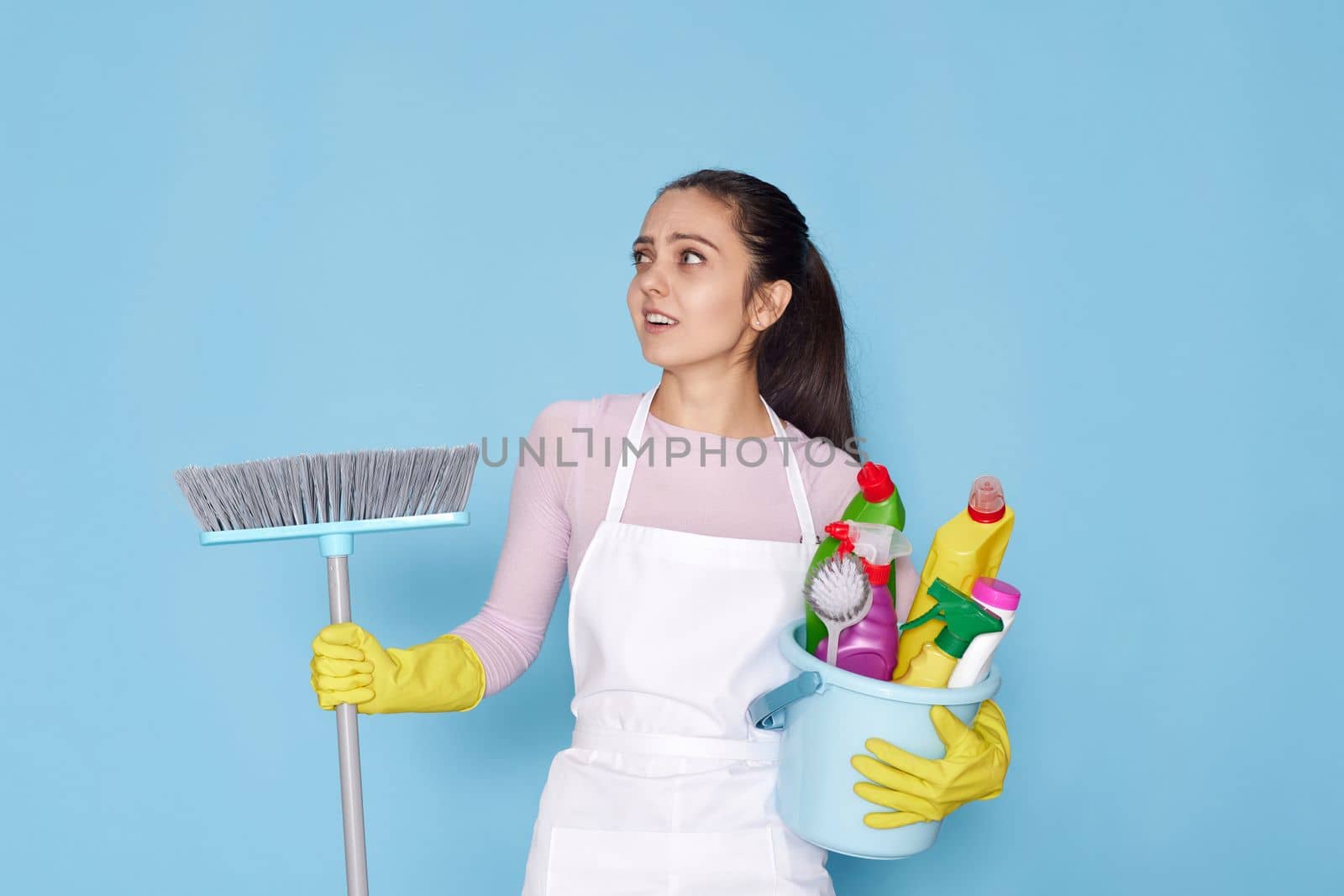 skeptic and nervous, frowning woman in gloves and cleaner apron holding bucket of detergents and broom on blue background.