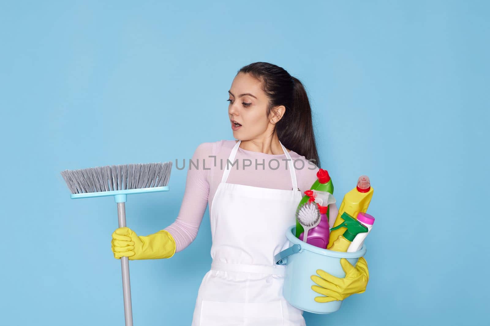 surprised young woman in rubber gloves and cleaner apron holding bucket of detergents and broom on blue background.