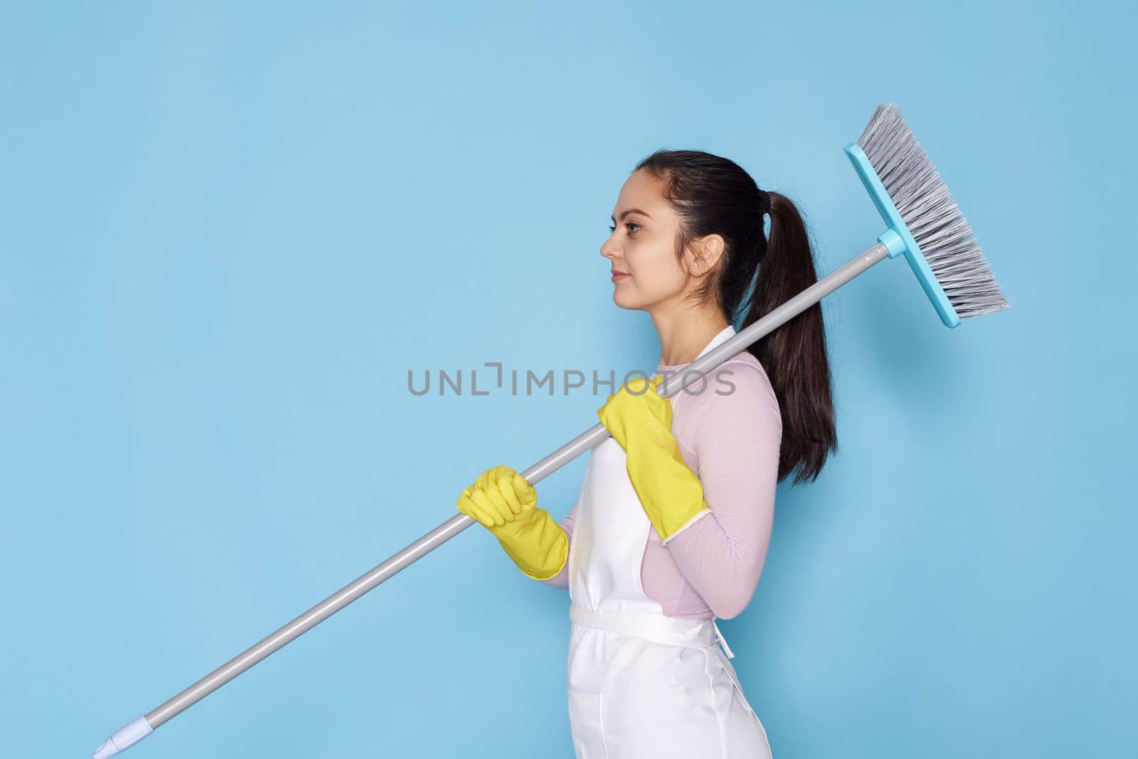 caucasian woman in rubber gloves and cleaner apron holding broom on blue background.