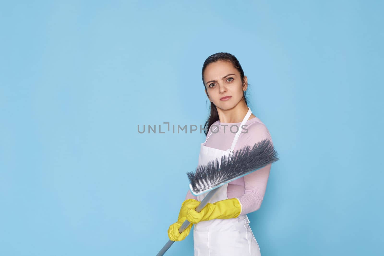 nervous, frowning woman in rubber gloves and cleaner apron with broom on blue background.