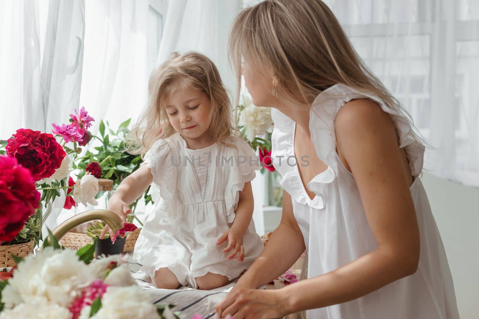 A little blonde girl with her mom on a kitchen countertop decorated with peonies. The concept of the relationship between mother and daughter. Spring atmosphere.