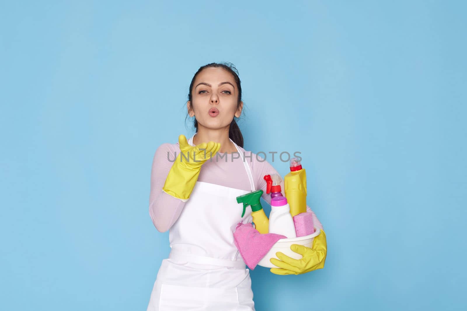 caucasian woman in gloves and cleaner apron send air kiss on blue background.