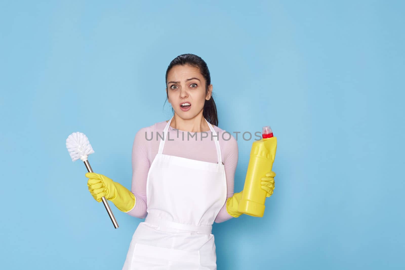 surprised nervous, frowning woman in yellow rubber gloves and cleaner apron holding toilet brush and bottle of detergent on blue background.