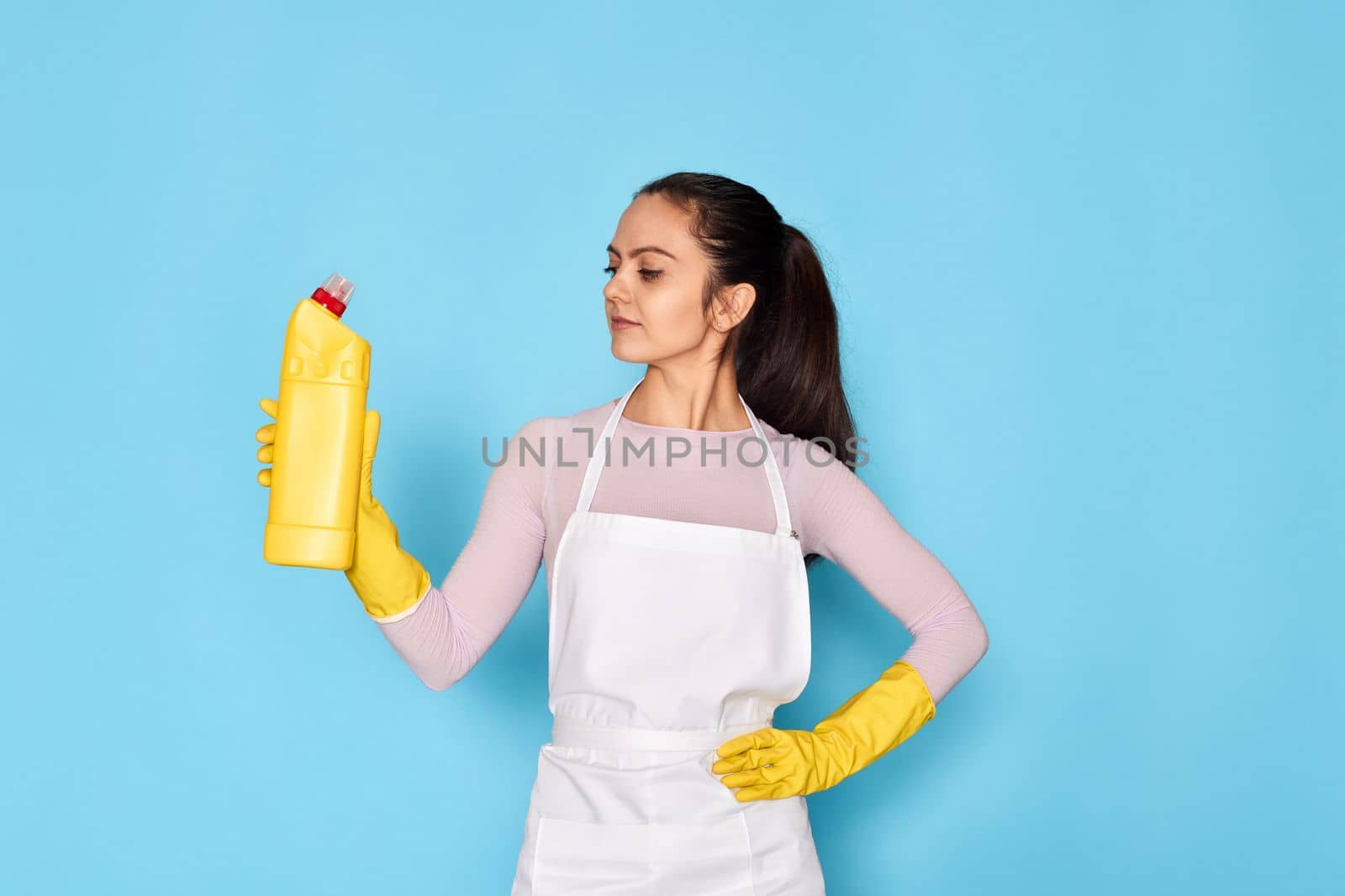 beautiful caucasian woman in yellow rubber gloves and cleaner apron holding bottle of detergent on blue background.