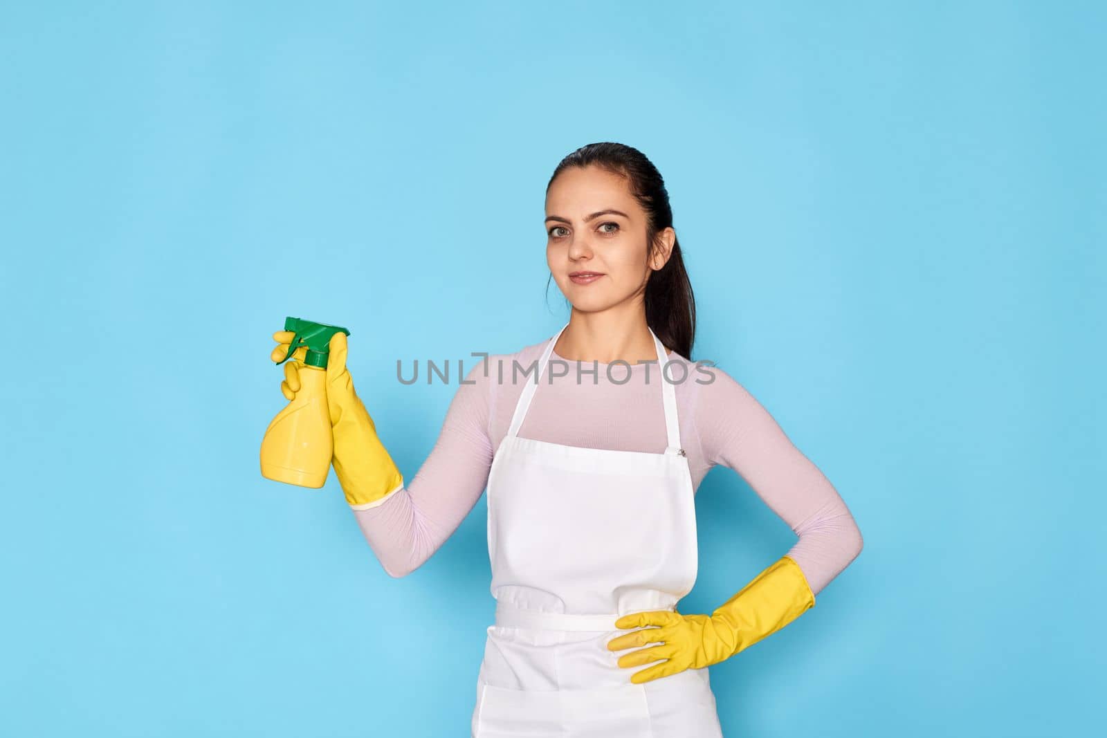 young confident woman in rubber gloves and cleaner apron holding detergent sprayer on blue background.