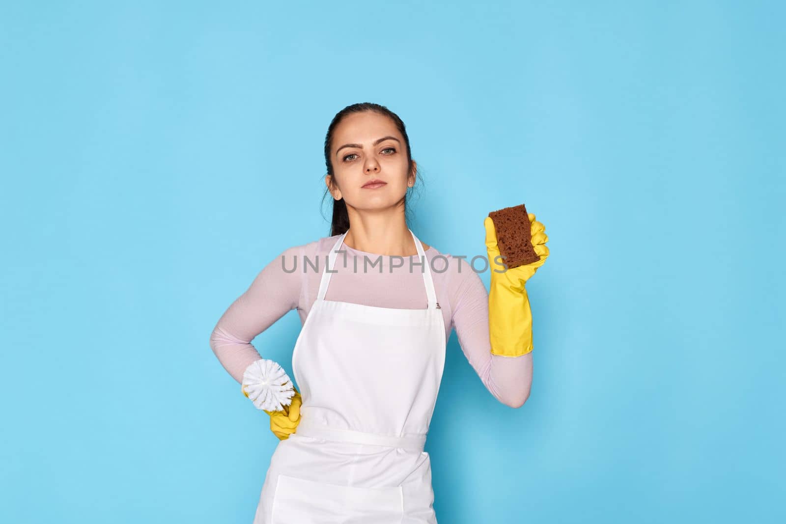 diligent woman in rubber gloves and cleaner apron cleaning with sponge on blue background.