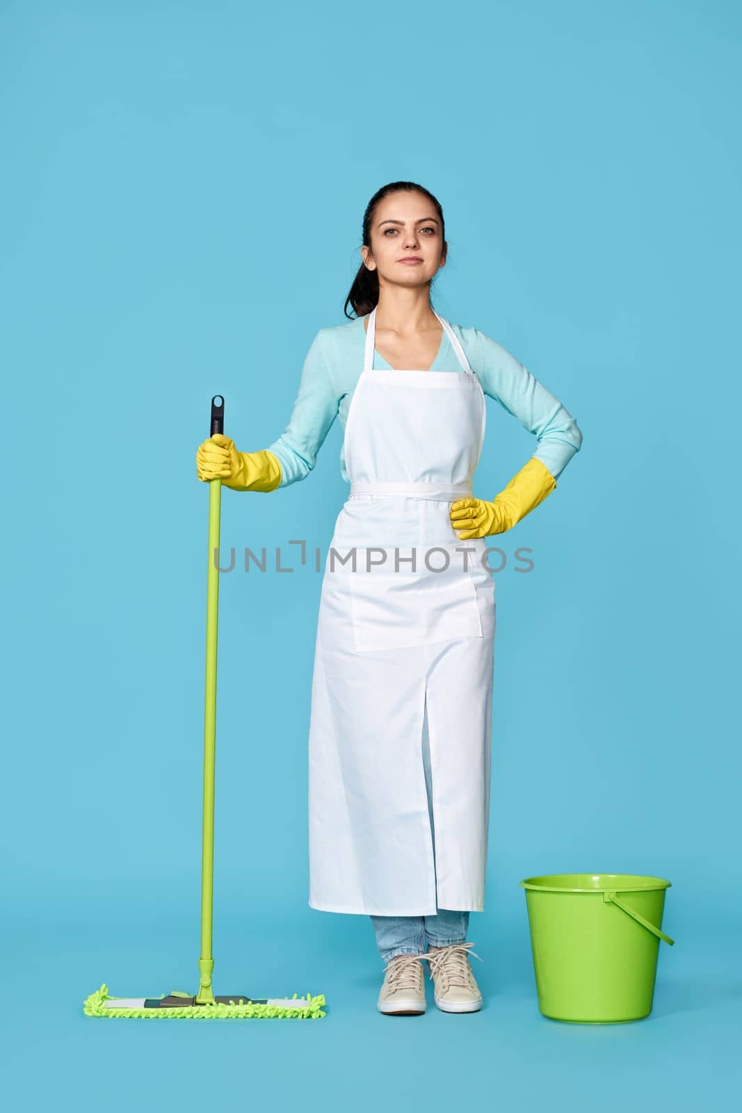 professional cleaner girl in rubber gloves and cleaner apron washing floor with mop on blue background. Full length