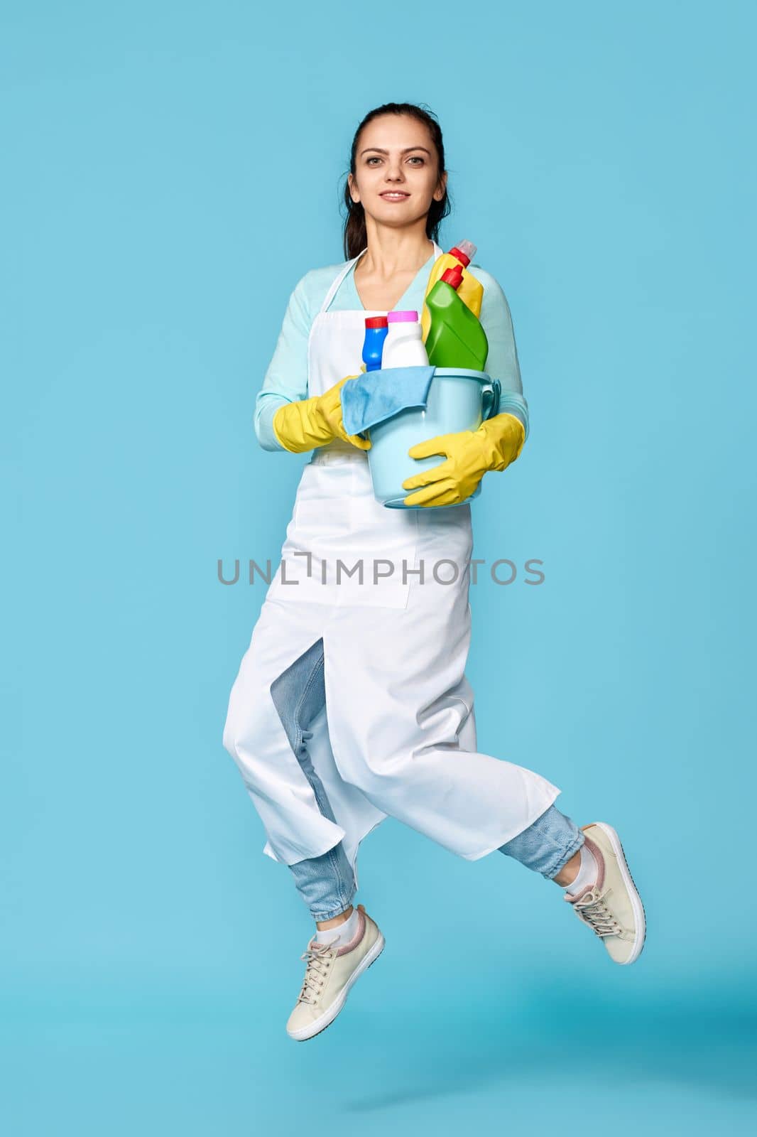 happy cheerful woman in rubber gloves and cleaner apron with cleaning supplies jumping on blue background. Full length