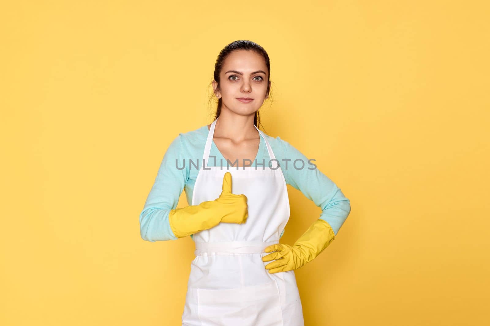 happy caucasian woman in rubber gloves and cleaner apron showing thumbs up gesture on yellow background. cleaning