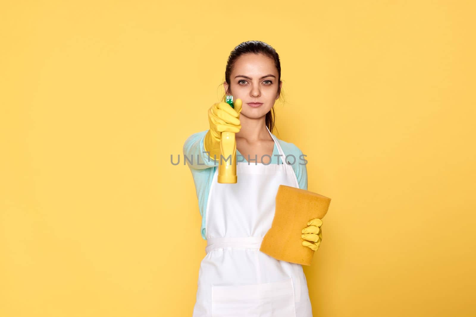 caucasian woman in yellow rubber gloves and cleaner apron with sponge and detergent sprayer on yellow background.