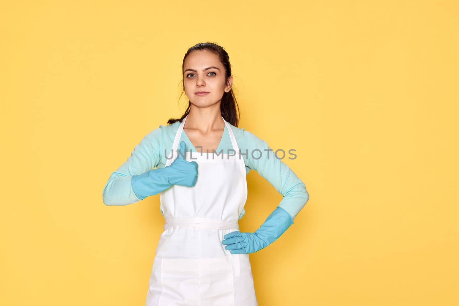 happy caucasian woman in blue rubber gloves and cleaner apron showing thumbs up gesture on yellow background. cleaning