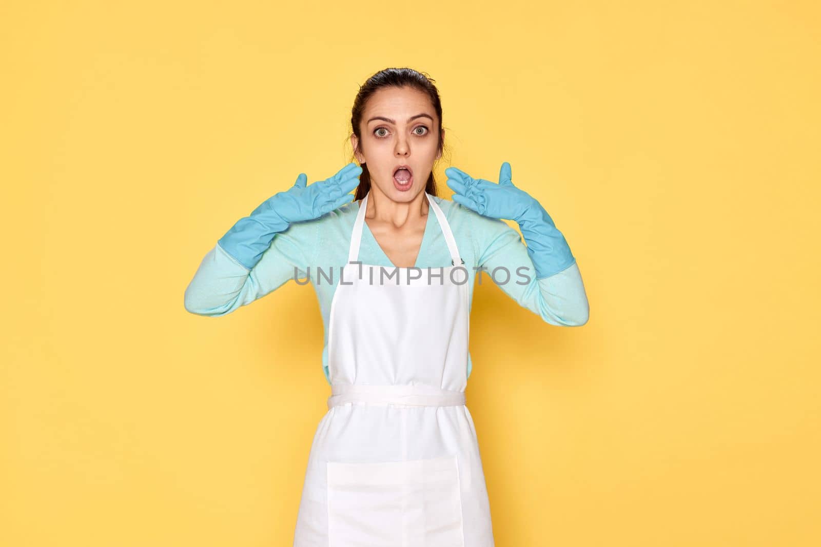 surprised young woman in blue rubber gloves and cleaner apron on yellow background.