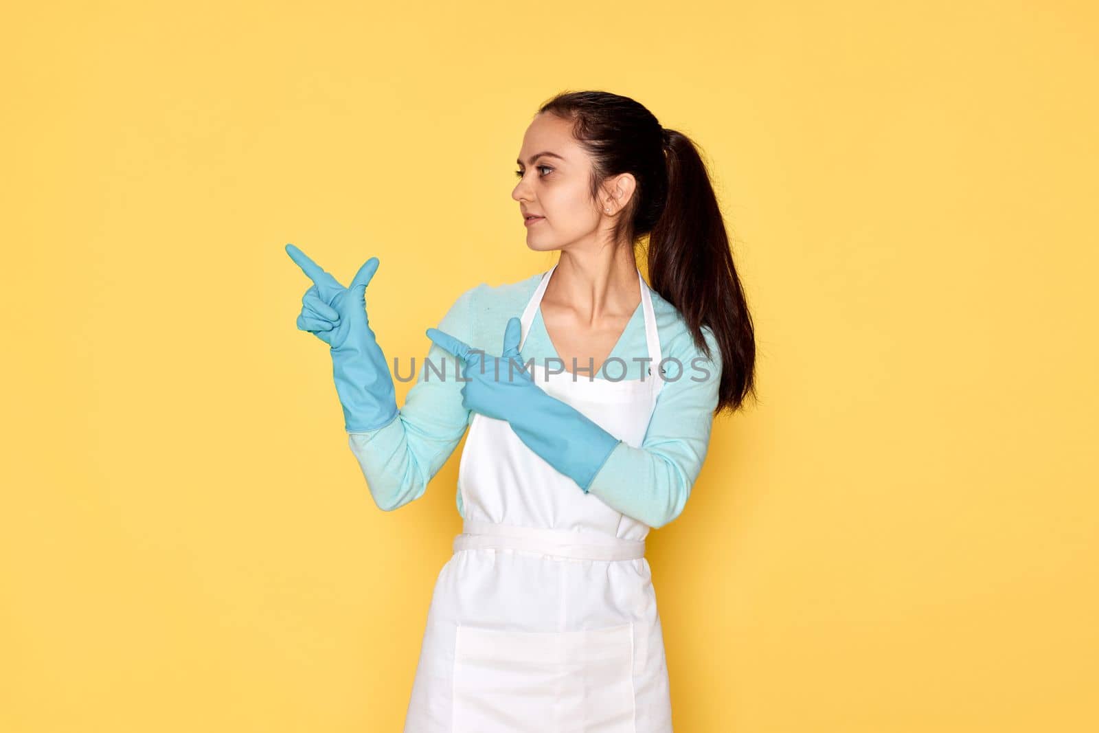 beautiful caucasian woman in rubber gloves pointing fingers aside at copy space on yellow background. cleaning