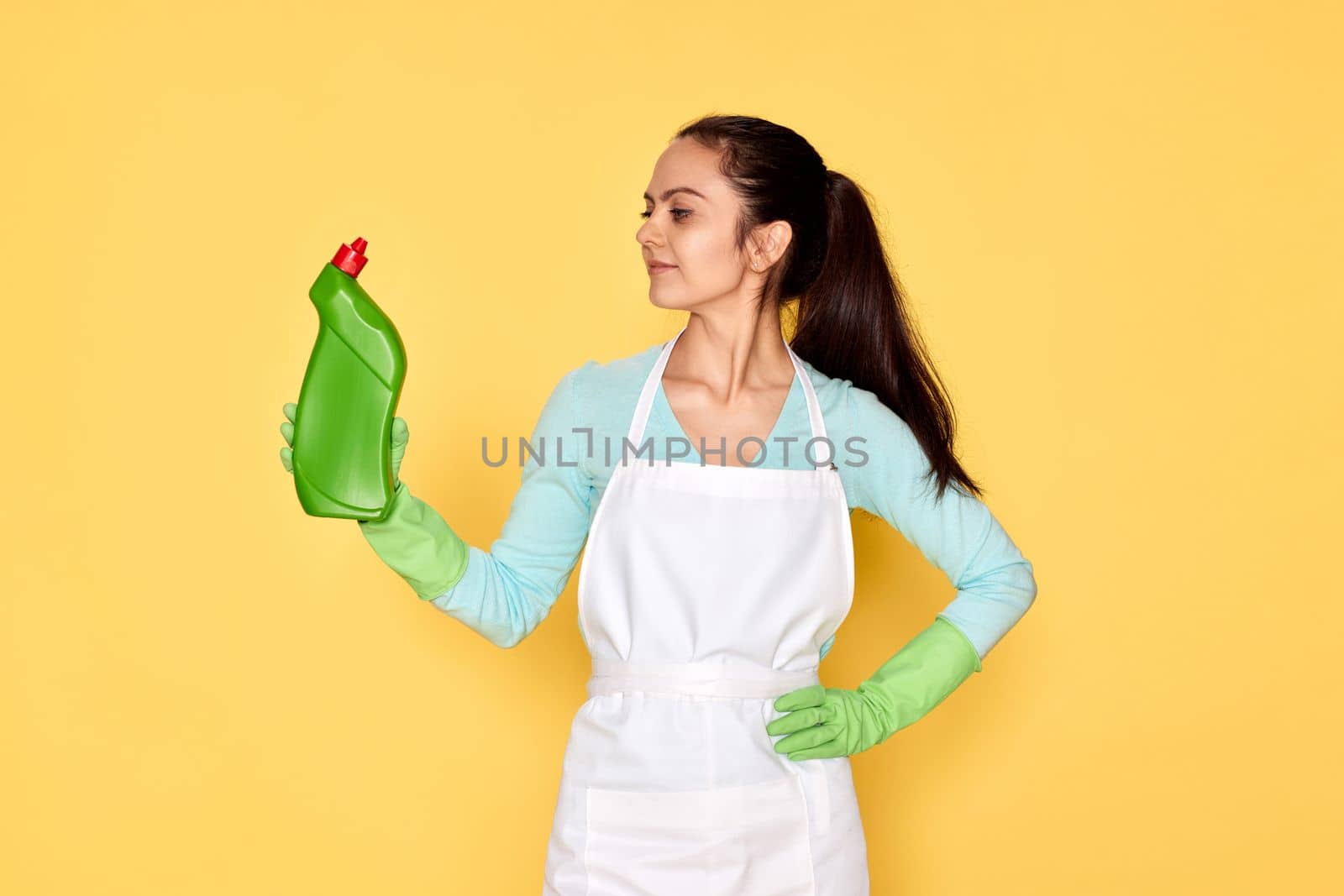 beautiful caucasian woman in rubber gloves and cleaner apron holding bottle of detergent on yellow background.