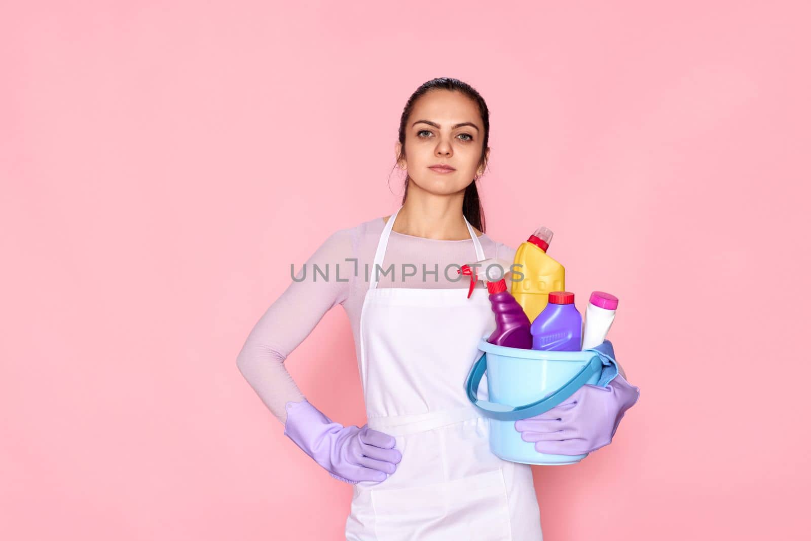 pretty caucasian woman in gloves holding bucket with cleaning supplies isolated on pink background.