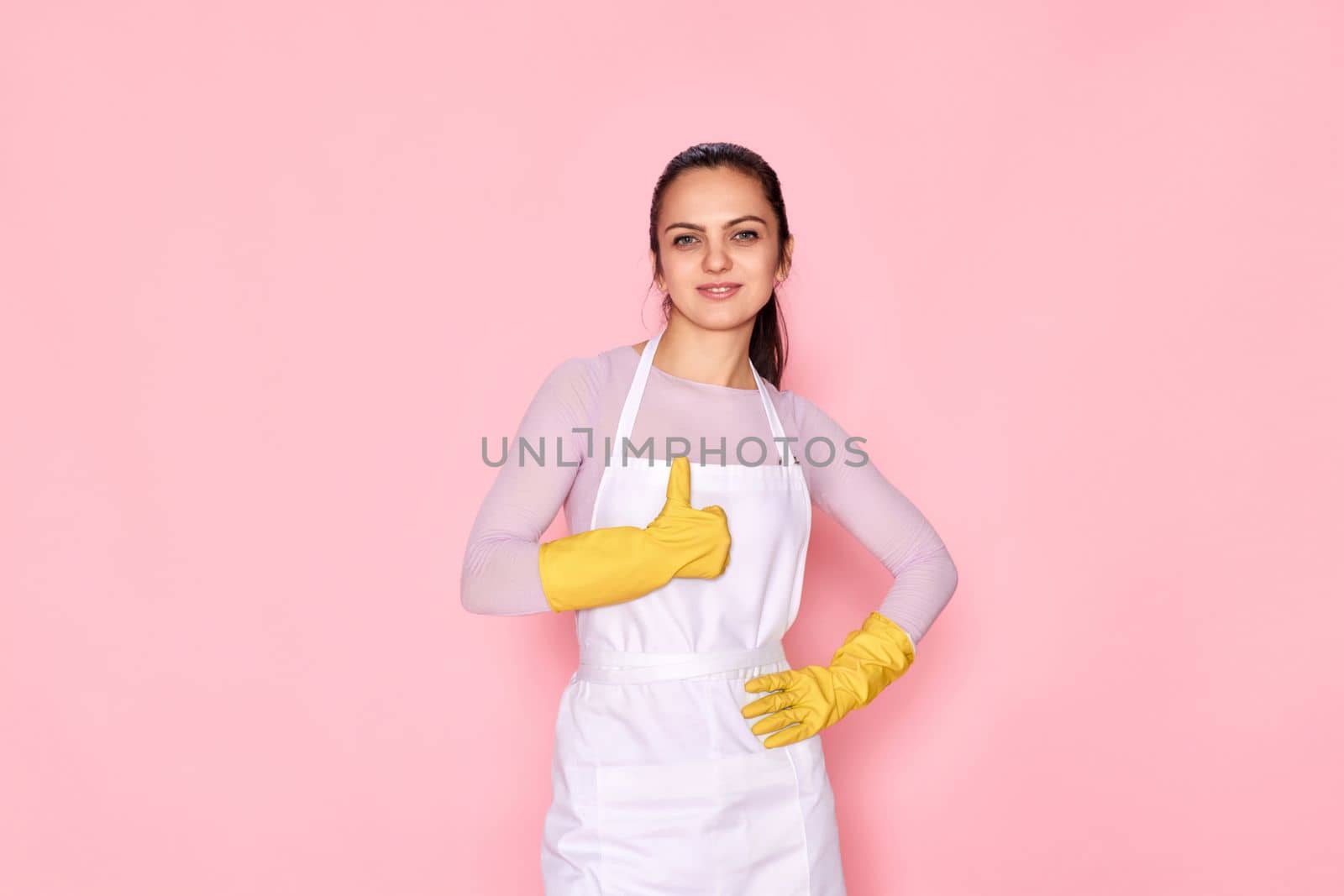 happy caucasian woman in yellow rubber gloves and cleaner apron showing thumbs up gesture on pink background. cleaning