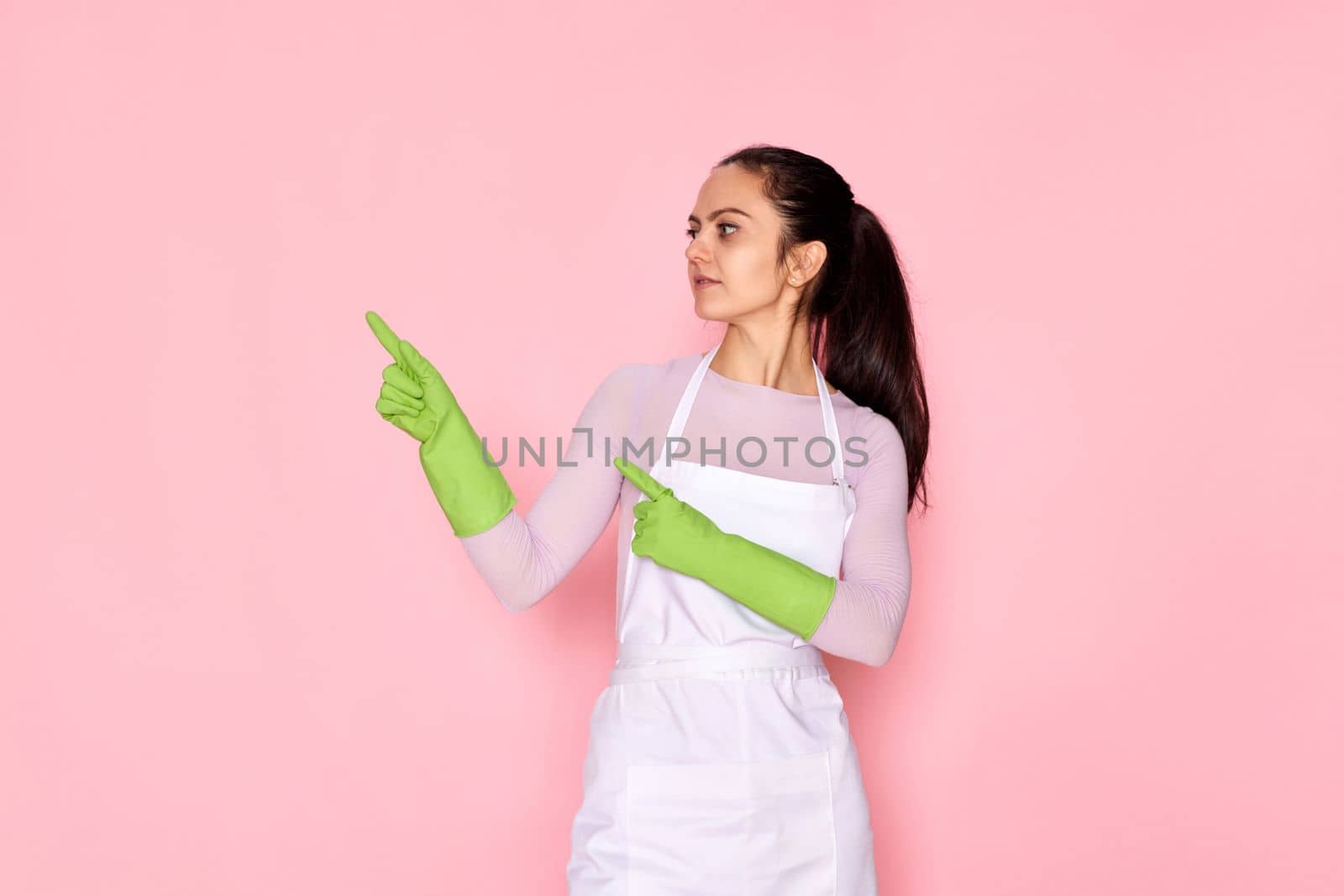 beautiful caucasian woman in rubber gloves pointing fingers aside at copy space on pink background. cleaning