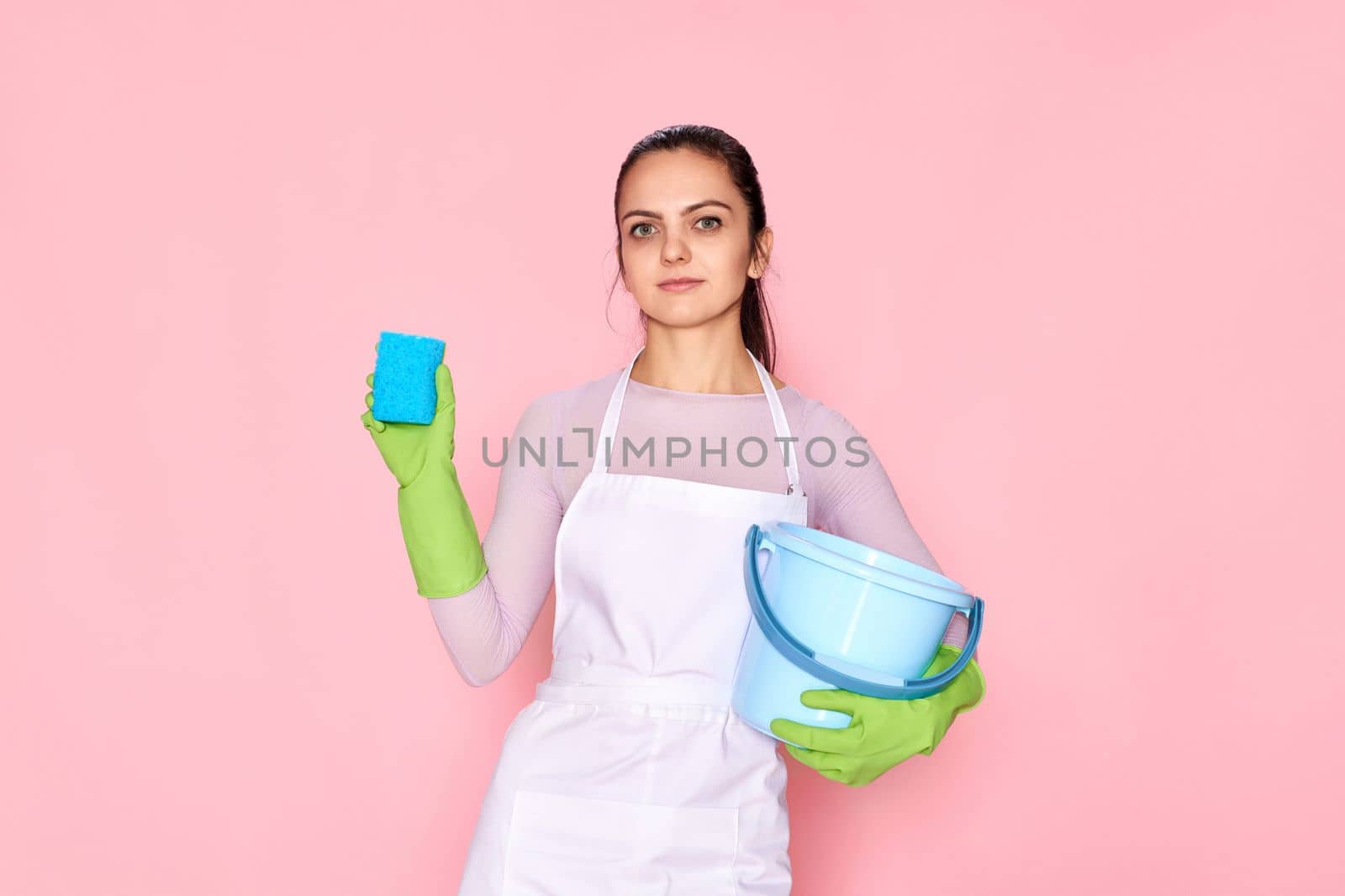 young confident woman in rubber gloves and cleaner apron cleaning with blue sponge and bucket on pink background.